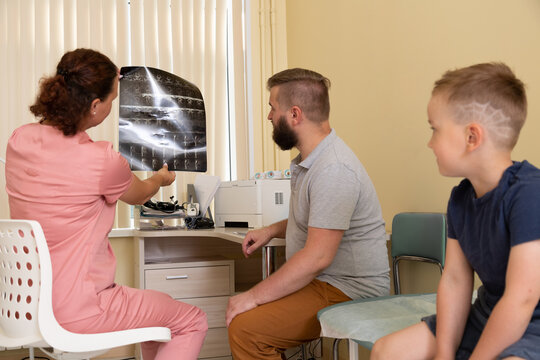 Horizontal photo. Woman doctor in pink uniform with patient, man with beard , boy, son looks at radiology X-ray film shot in hospital office of private clinic. Concept of health care, treatment, work