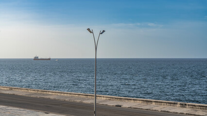 A ship in the blue ocean. Ripples on calm water. Azure sky, clouds. In the foreground is the Malecon embankment, a street lamp. Cuba. Havana. 