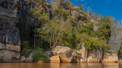 Trees grow on the steep, eroded coastal cliffs. Reflection on the surface of a red-brown river. Blue sky, clouds. Madagascar. Manambolo river.