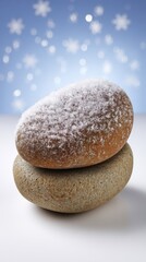 Two natural, smooth zen stones stacked one on top of the other, with the upper rock dusted in snow, set against a shimmering blue winter bokeh background.