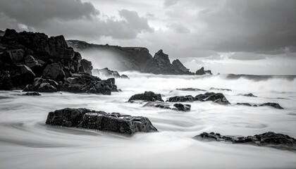A dramatic black and white long exposure captures the raw power of ocean waves crashing against jagged coastal rocks under a moody, cloudy sky.