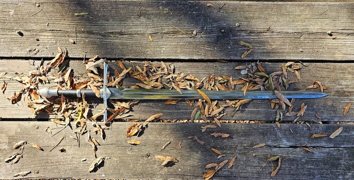Sword on wooden surface partly covered by autumn leaves and sunlight and shadows - Powered by Adobe