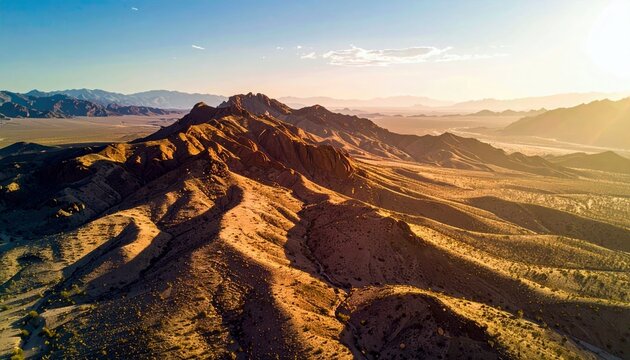 An aerial view of a vast desert landscape at sunset, showcasing rugged mountains bathed in warm golden light. - Powered by Adobe