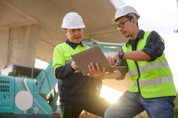 Civil engineers reviewing construction plans on laptop while supervising road construction work, coordinating field operations and inspecting elevated highway infrastructure.