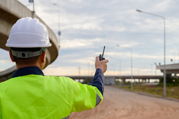 Construction engineer supervising road construction using walkie talkie, coordinating field work and inspecting elevated highway infrastructure.