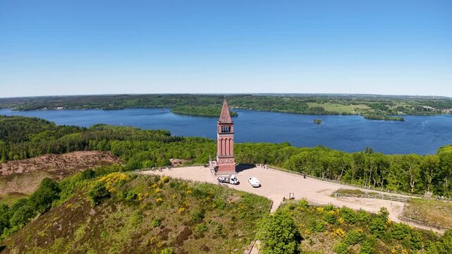 A drone soars high above the Himmelbjerget tower perched on a peninsula, revealing a panoramic view of the surrounding lake and lush green forest in Denmark.