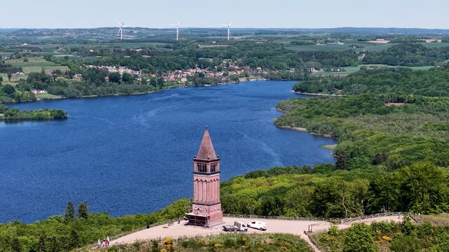 A high-flying drone captures a wide shot of the Himmelbjerget tower on a hill beside a large blue lake with surrounding forests in Denmark.