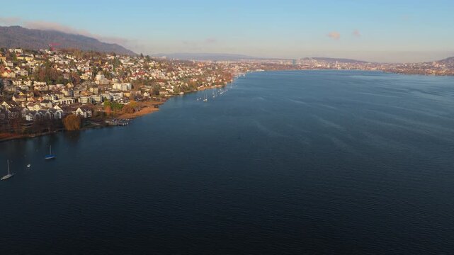 Drone captures wide aerial view from the middle of Lake Zurich. Clear daylight reveals the shoreline of Kilchberg and Zurich city in the distance across calm blue water.