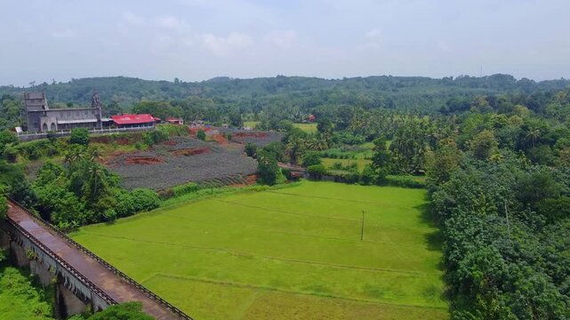 Wide aerial view of an historic stone bridge aqueduct over a valley, showcasing bright green paddy fields and a pineapple plantation leading up to a large Christian church in Kerala, India.