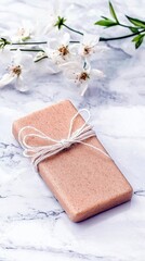 A single bar of soap, wrapped in brown paper and tied with twine, sits on a marble surface with white blossoms and green leaves.