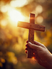 Hand holding a wooden Christian cross against a warm glowing sunrise with bokeh background symbolizing faith and spirituality