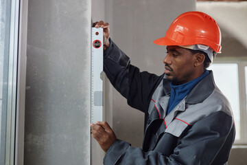 Black middle aged man wearing hard hat using spirit level tool measuring wall surface indoors, focusing on accuracy during construction or renovation project, standing near window