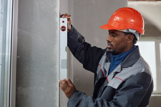 Black middle aged man wearing hard hat using spirit level tool measuring wall surface indoors, focusing on accuracy during construction or renovation project, standing near window