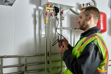 Caucasian middle aged man inspecting heating system pipes and holding electrical wires while working in utility room, wearing safety vest and focusing on maintenance task