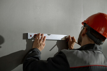 Middle aged Caucasian man wearing hard hat using spirit level and pencil marking measurements on wall, focusing on precise alignment during construction or renovation work