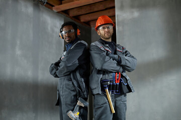 Two young adult men, one Black and one Caucasian, standing back to back with arms crossed, wearing protective workwear and safety gear, posing confidently at construction site