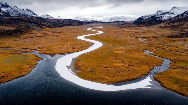 A wide, winding river flows through a barren, golden-brown landscape towards snow-capped mountains under a dramatic, cloudy sky.