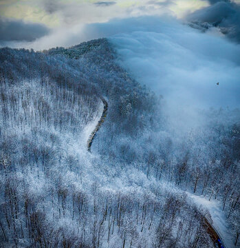Aerial view of a winding road cuts through a snow-laden forest, shrouded in mist and clouds along Asalem - Khalkhal Road, Gilan Province, Iran. 