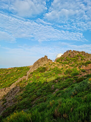 Scenic landscape with rocky hills covered in green vegetation under a bright blue sky with soft, scattered clouds. Natural outdoor scenery, summer mountain view, peaceful nature background.