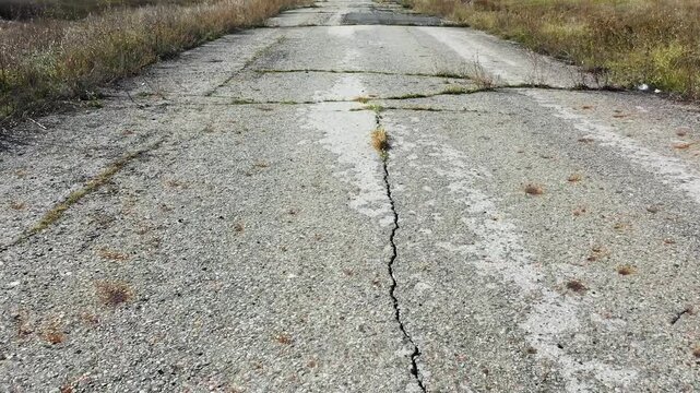 Aerial 4K footage of an abandoned asphalt road. Grass breaks through and grows from cracks in the asphalt. Damaged cracked road path with weeds growing Nature takes its toll.