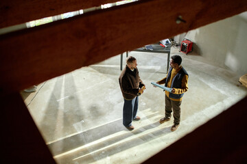 Caucasian middle aged man and Black middle aged man standing in unfinished room discussing construction project, holding blueprint, sunlight streaming through windows onto concrete floor