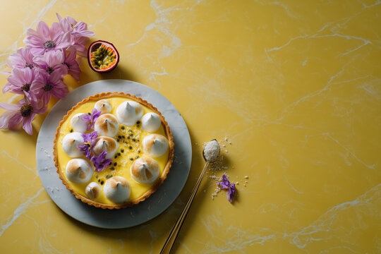 Delicious passion fruit tart with meringue kisses, delicate flowers, and golden spoon on vibrant yellow marble for a tempting dessert indulgence