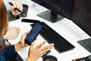 Cropped pointing hand with pen in businessman, checking with holding smartphone by woman investor online website program application with dynamic stock market at home office at back side. Infobahn.