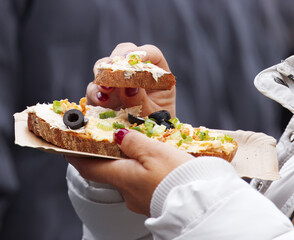 Visitor at Prague Naplavka market enjoys autumn open-faced sandwich topped with creamy spread, olives, spring onions and crunchy garnish, capturing cozy seasonal vibe of this lively riverside market.