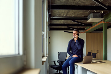 Professional Man on Phone in Modern Office With Laptop at Desk and Large Windows