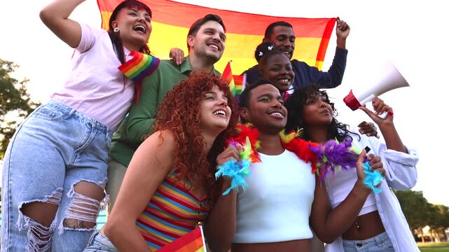 Group of happy young multiracial friends standing together in a park, holding a large rainbow flag and celebrating gay pride with smiles, while a woman shouts into a megaphone