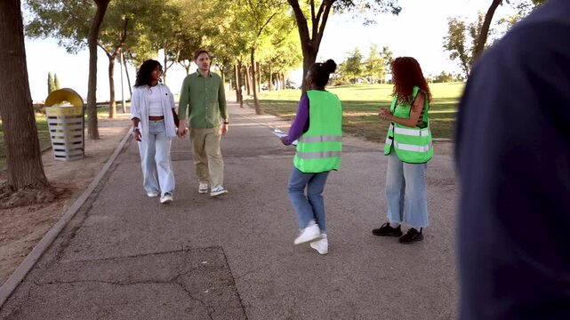 Two young diverse charity workers wearing green reflective vests stop people on a park path to talk and ask for support, engaging passersby for their non profit organization's cause