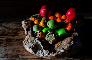 Mixed fruit including apples, pears, persimmons, oranges and pomegranates arranged in a textured wooden bowl with strong side-light and deep shadows