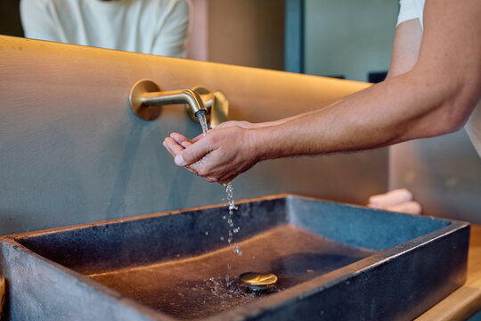 Person washing hands under running water for hygiene and cleanliness. Water flowing from a wall-mounted faucet into a modern sink
