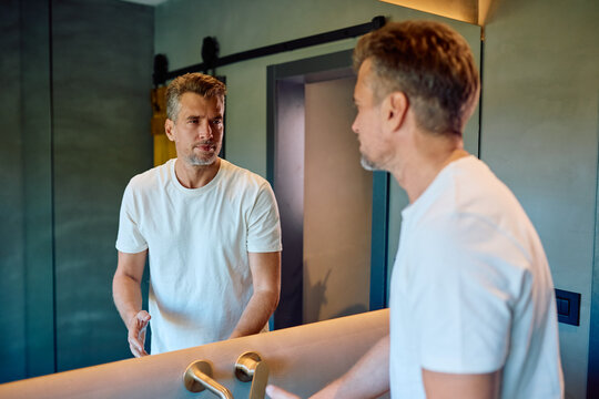 Middle-aged man standing in front of a mirror, looking at his reflection and introspecting. Thinking about aging and self-acceptance