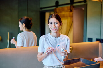 Young woman standing in a modern bathroom, smiling at the camera while holding a toothbrush and tube of toothpaste