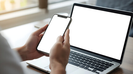 A person is seated at a desk, holding a smartphone and facing an open laptop, both displaying blank white screens. The indoor workspace is illuminated by natural light from a window.