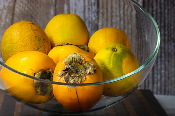 glass bowl with fresh persimmons and oranges on wooden surface