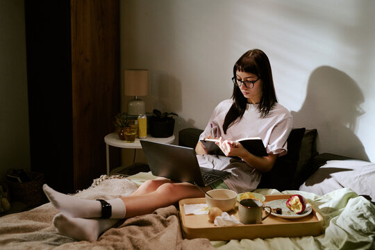 Young adult biracial woman sitting on bed working on laptop and writing in notebook with electronic ankle monitor visible on leg, breakfast tray with food and drink nearby - Powered by Adobe