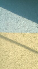 Textured split wall featuring pastel blue and yellow surfaces intersected by soft diagonal shadows creating a minimalist abstract pattern