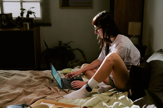 Young adult biracial woman sitting on bed using laptop with electronic ankle monitor visible on leg, staying at home in casual setting working or browsing internet during house arrest
