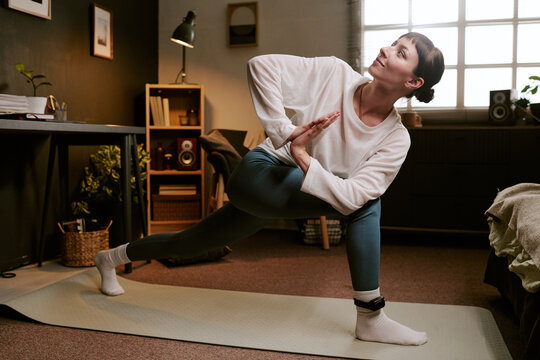 biracial young adult woman practicing yoga at home wearing ankle monitor, stretching on exercise mat in living room, focusing on fitness and rehabilitation during house arrest - Powered by Adobe