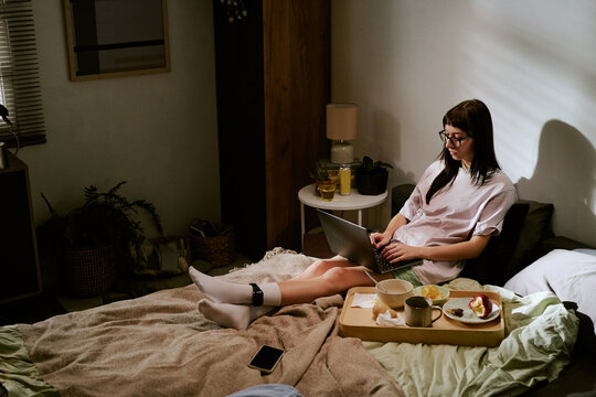 biracial young adult woman sitting on bed working on laptop with ankle monitor visible on leg, staying at home with food tray nearby, electronic device on blanket, casual indoor setting
