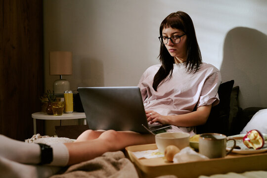 Young adult biracial woman sitting on bed using laptop with electronic ankle monitor visible on leg, staying at home under house arrest focusing on screen with breakfast tray nearby