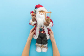 Young adult woman hands holding and showing Santa Claus toy on light blue table background. Pastel...