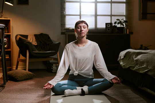 Young adult biracial woman sitting cross legged on floor meditating with eyes closed, wearing ankle monitor in home setting sunlight streaming through window in background