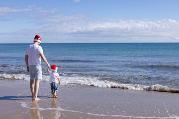 Father and his little son wearing Santa hats walking on a beach. Christmas concept.