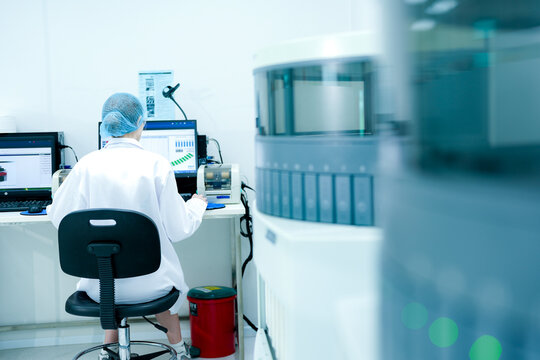 A close up of a female scientist in a hairnet and safety glasses using a computer. She is monitoring data or results from an automated analyzer in a modern laboratory.