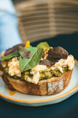 Gourmet close-up of a rustic sourdough toast layered with creamy avocado and scrambled eggs, topped with fresh greens
