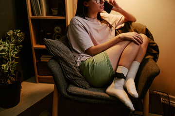 Obraz premium Young adult biracial woman sitting on chair at home wearing electronic ankle monitor, talking on phone, resting legs on seat, partially visible face with long hair and glasses