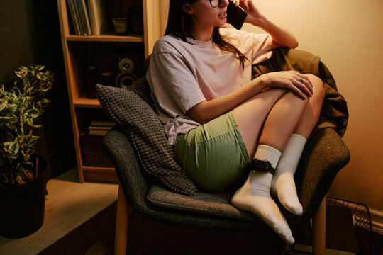 Young adult biracial woman sitting on chair at home wearing electronic ankle monitor, talking on phone, resting legs on seat, partially visible face with long hair and glasses